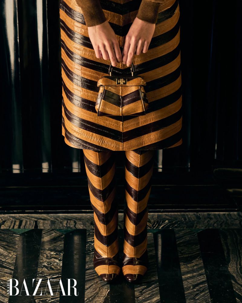 Woman in striped skirt and pants holding a purse with hands, posing indoors on a striped floor with dark background; fashion advertisement.