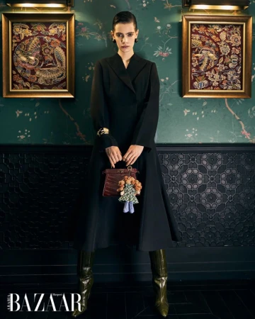 Model in a structured black coat dress and olive green boots holding a handbag with a doll charm before green floral wallpaper.