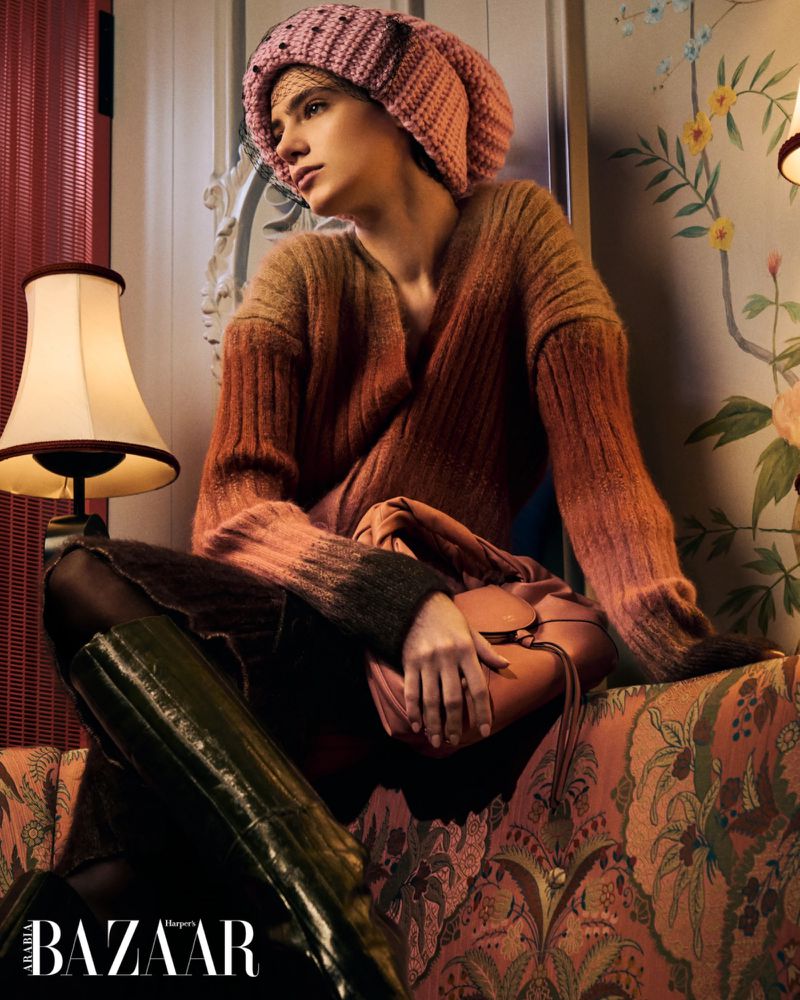 Fashionable woman in sweater, knit hat, and shiny boots poses indoors against patterned wallpaper with a handbag and lamp.
