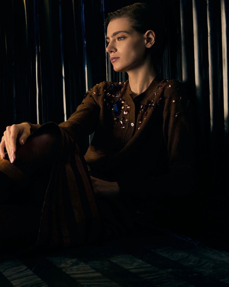 Portrait of a stylish young woman with dark hair wearing a brown embellished cardigan and trousers against a dark corrugated background.