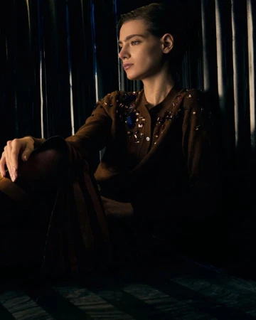 Portrait of a stylish young woman with dark hair wearing a brown embellished cardigan and trousers against a dark corrugated background.