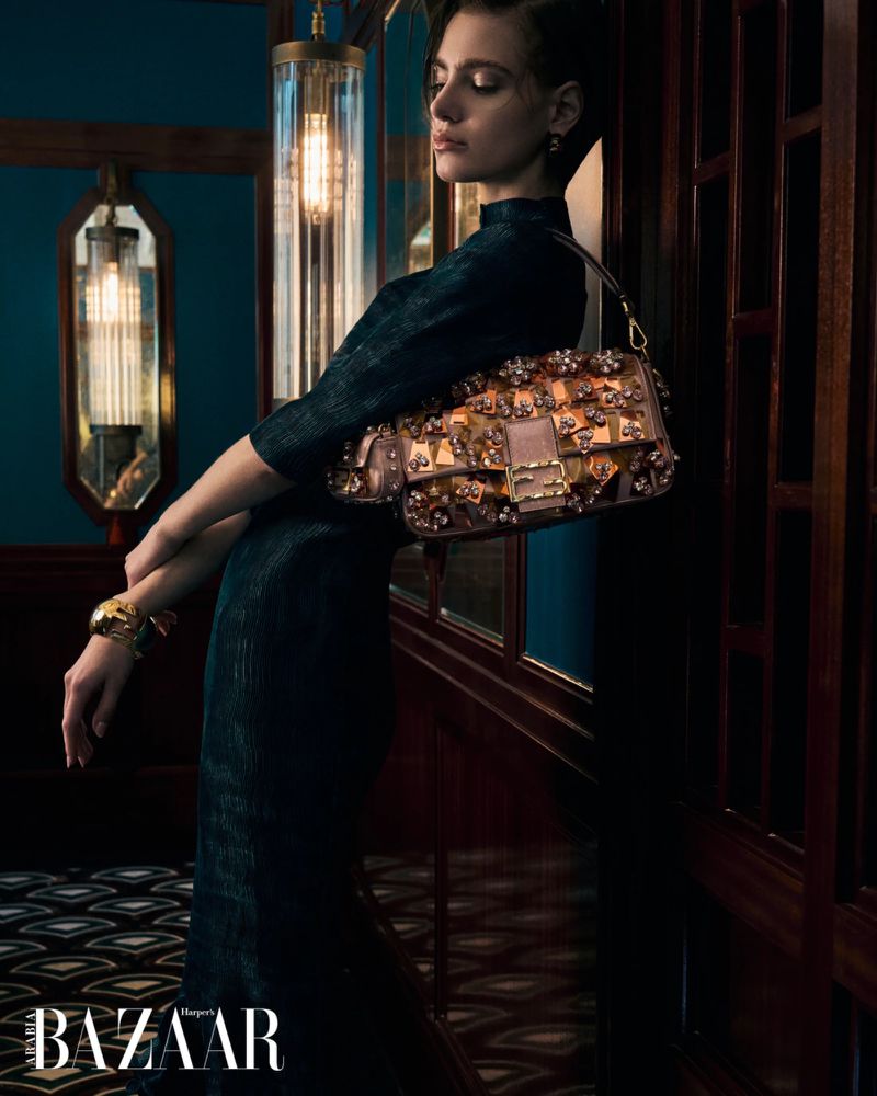 Adult woman in a black dress poses indoors with a brown handbag adorned with metallic and floral details, showcasing fashion and style.