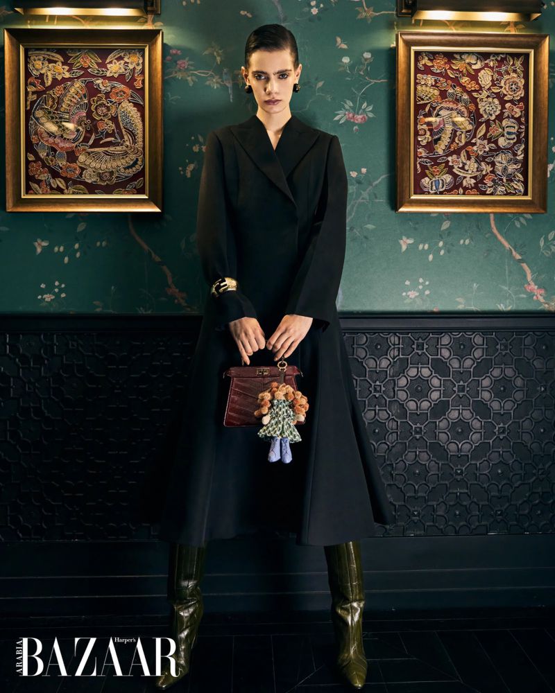 Model in a structured black coat dress and olive green boots holding a handbag with a doll charm before green floral wallpaper.
