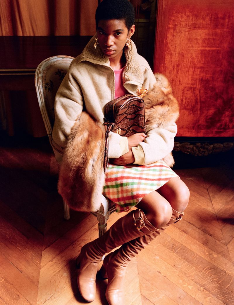 Fashion portrait of an adult woman with an afro, wearing a pink top, plaid skirt, beige jacket, fur stole, tan boots, and holding a handbag.