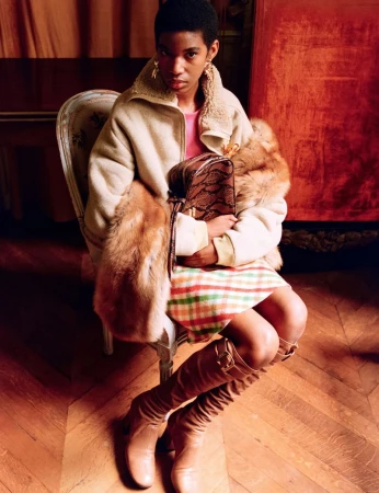 Fashion portrait of an adult woman with an afro, wearing a pink top, plaid skirt, beige jacket, fur stole, tan boots, and holding a handbag.