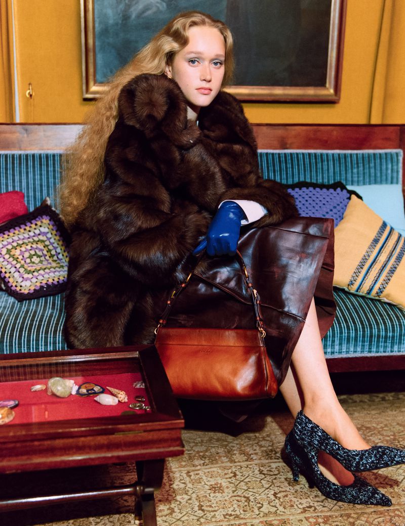 A blonde woman in a fur coat sits on a vintage couch with a handbag, styled with blue gloves and patterned heels, captured in a medium shot.
