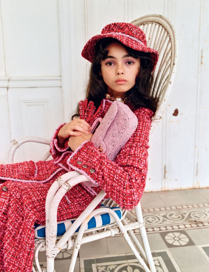 Young girl in red tweed hat and suit holds a pink purse while seated in a white wicker chair with blue striped cushion indoors.