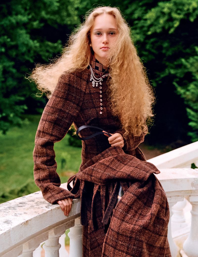 A young woman with curly blond hair poses on a white stone balcony, wearing a brown plaid high-necked coat with a black belt and pendant necklace.