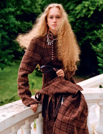 A young woman with curly blond hair poses on a white stone balcony, wearing a brown plaid high-necked coat with a black belt and pendant necklace.