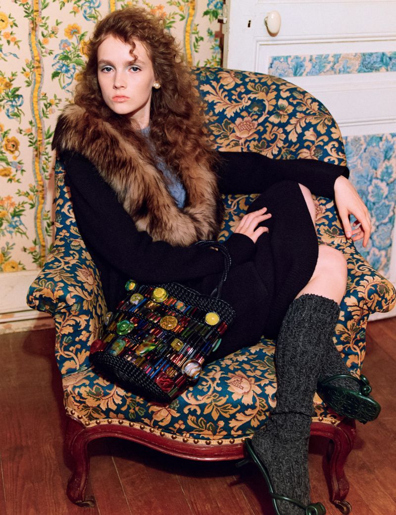 Portrait of a woman with auburn hair in a chair, wearing a fur collar and holding a colorful beaded purse, vintage wallpaper behind.