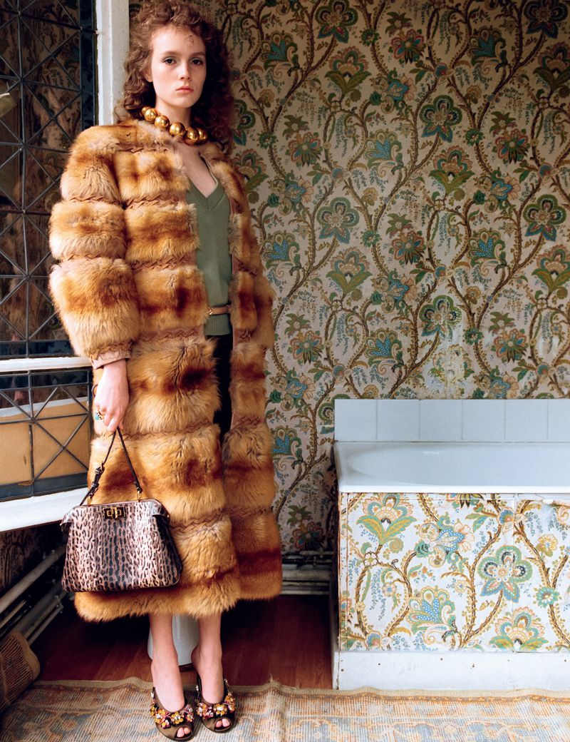 A model poses in a fur coat with a handbag indoors against wallpaper, showcasing a fashion aesthetic with a blend of luxury and vintage.