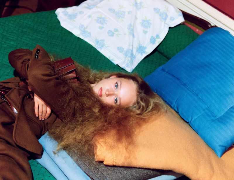 Young woman with long hair lies on bed with blue and brown pillows and green blanket, wearing a brown coat with belt.