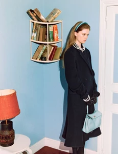 Fashion model wearing a long black coat, pearl necklaces, and a blue headband holding a light blue purse next to a corner bookshelf.