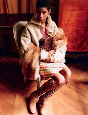 Seated woman wearing a beige fleece jacket, plaid skirt, and tan knee-high boots holding a brown snakeskin bag on an armchair.