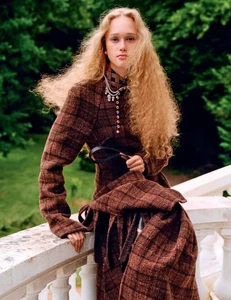 Model with long curly blonde hair wearing a brown plaid coat leaning against a white stone balustrade
