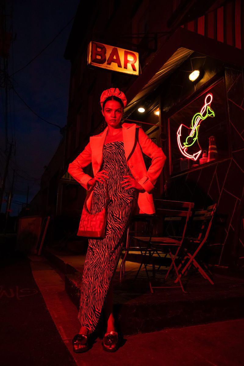 Woman in zebra-print jumpsuit and red blazer stands outside a bar with neon sign under the night sky, illuminated by red lighting.