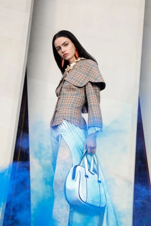 A solitary fashion model strikes a pose in a grey and brown plaid jacket dress, carrying a white handbag in a studio setting.