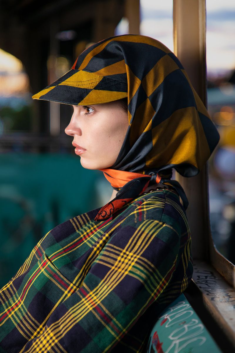 Profile portrait of a woman with a black and gold checkered headscarf and plaid shirt against an urban background with graffiti.