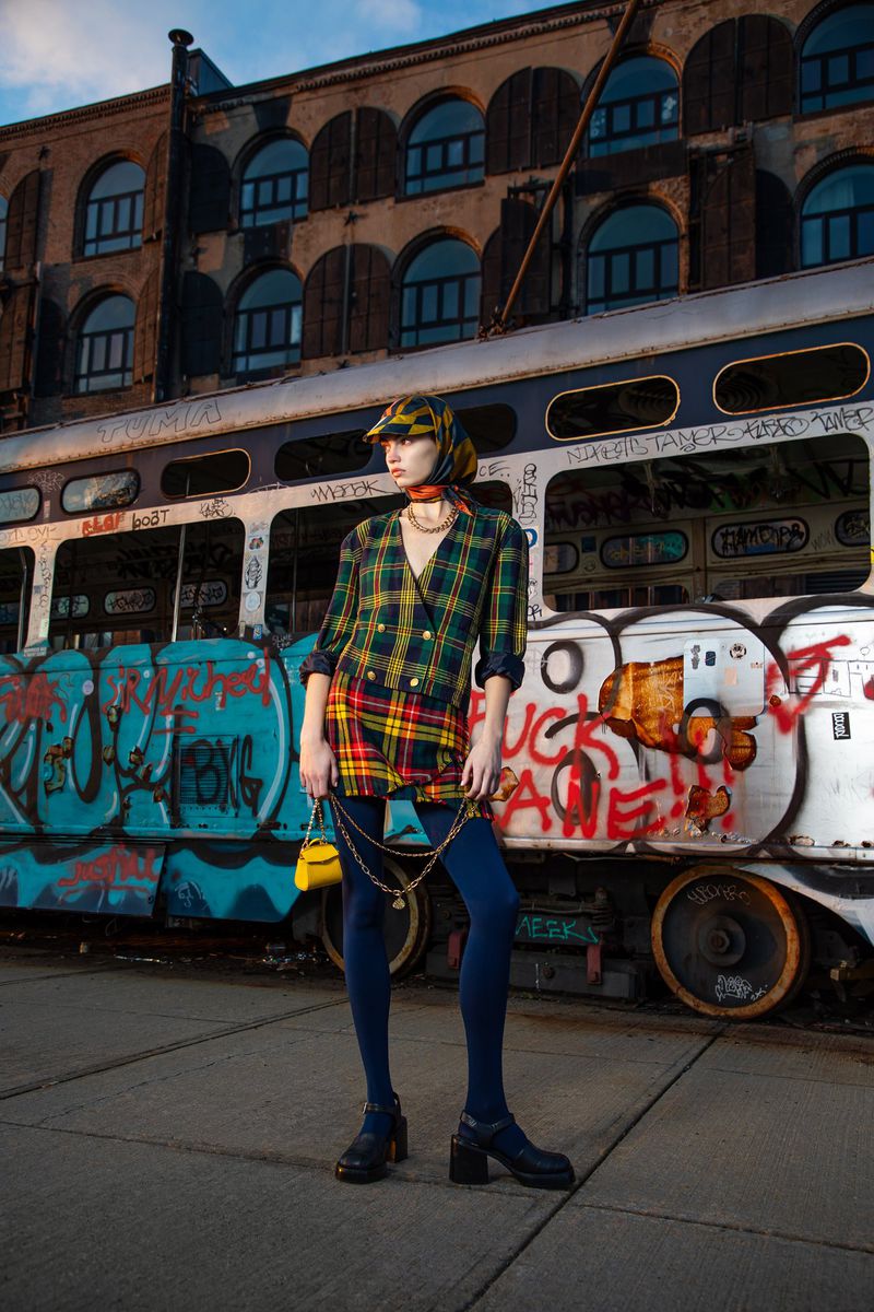 Fashionable woman in patterned suit poses in front of vintage trolley with graffiti and brick building backdrop. Urban editorial.