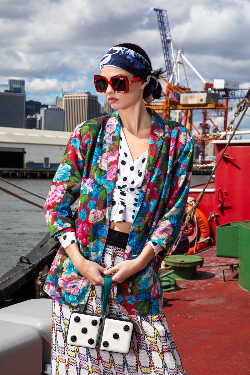 A woman in a floral jacket and geometric print skirt holds a dice-shaped purse with a cityscape and cloudy sky in the background.