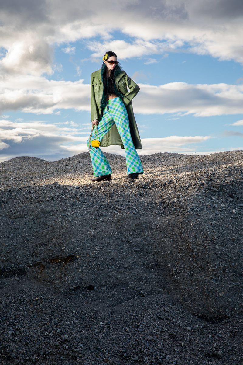 Outdoor fashion shot: a stylish woman in a green coat and plaid pants poses confidently on a rocky surface under a cloudy sky.