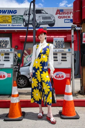 A woman wearing a floral jumpsuit and a red hat poses in front of a vintage gas station under a bright blue sky with a few clouds.