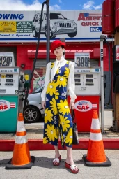 Model wearing blue and yellow floral overalls standing at a gas station with two orange traffic cones in foreground