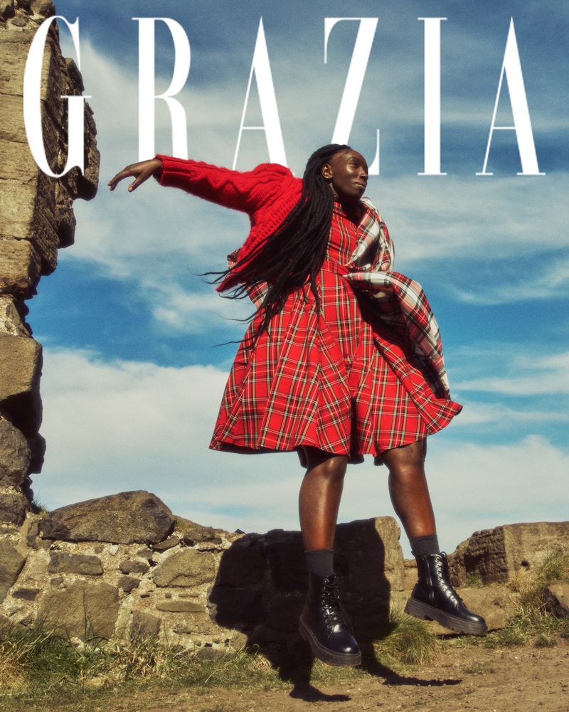 Fashion editorial shot of a black woman outdoors, wearing a red plaid dress, black boots, and braided hair, jumping near stone ruins.