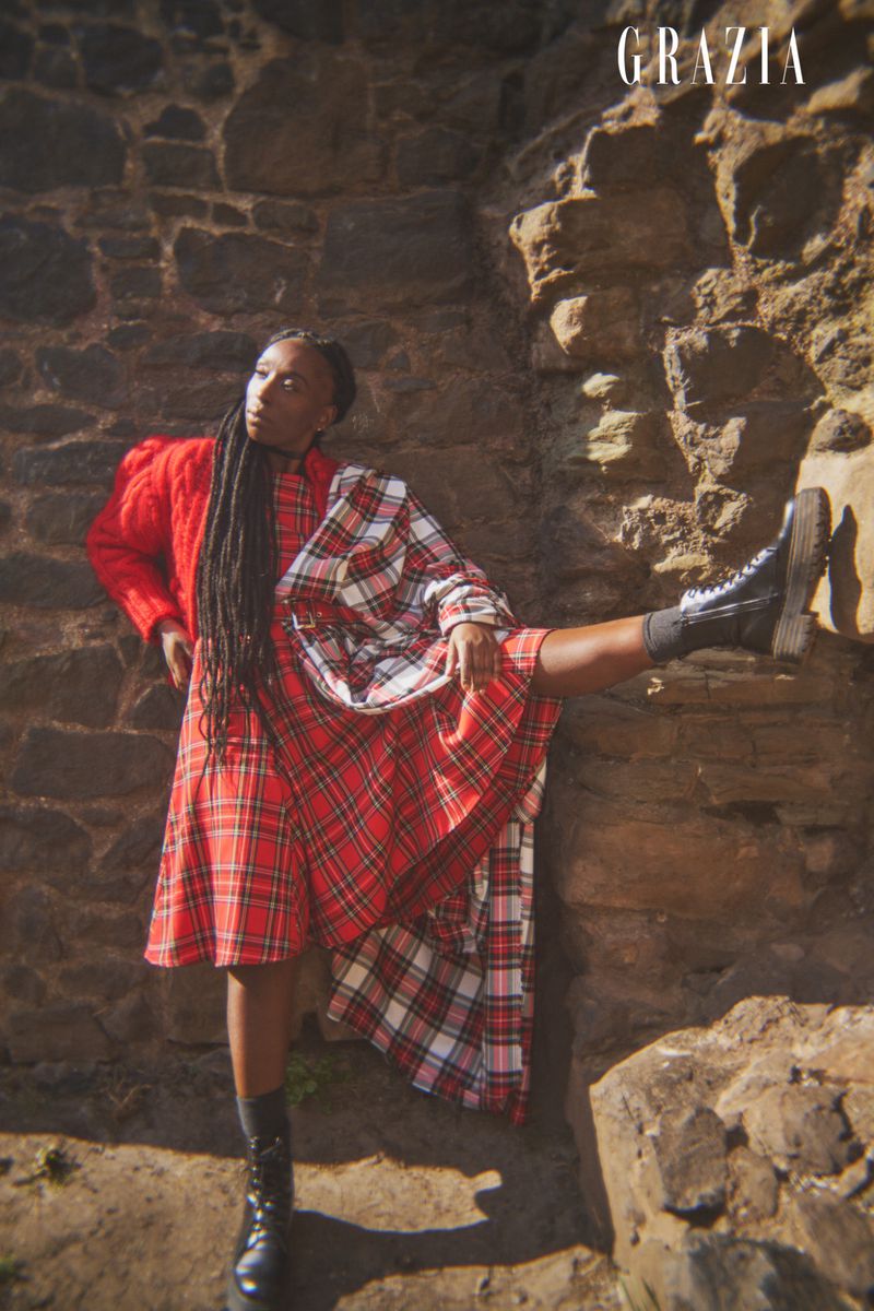 A woman in a red plaid dress and black boots poses against a weathered stone wall with the Grazia magazine logo visible.