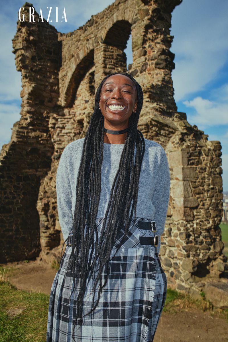 Smiling African-American woman in plaid skirt stands before ancient stone ruins under a blue sky with light clouds.