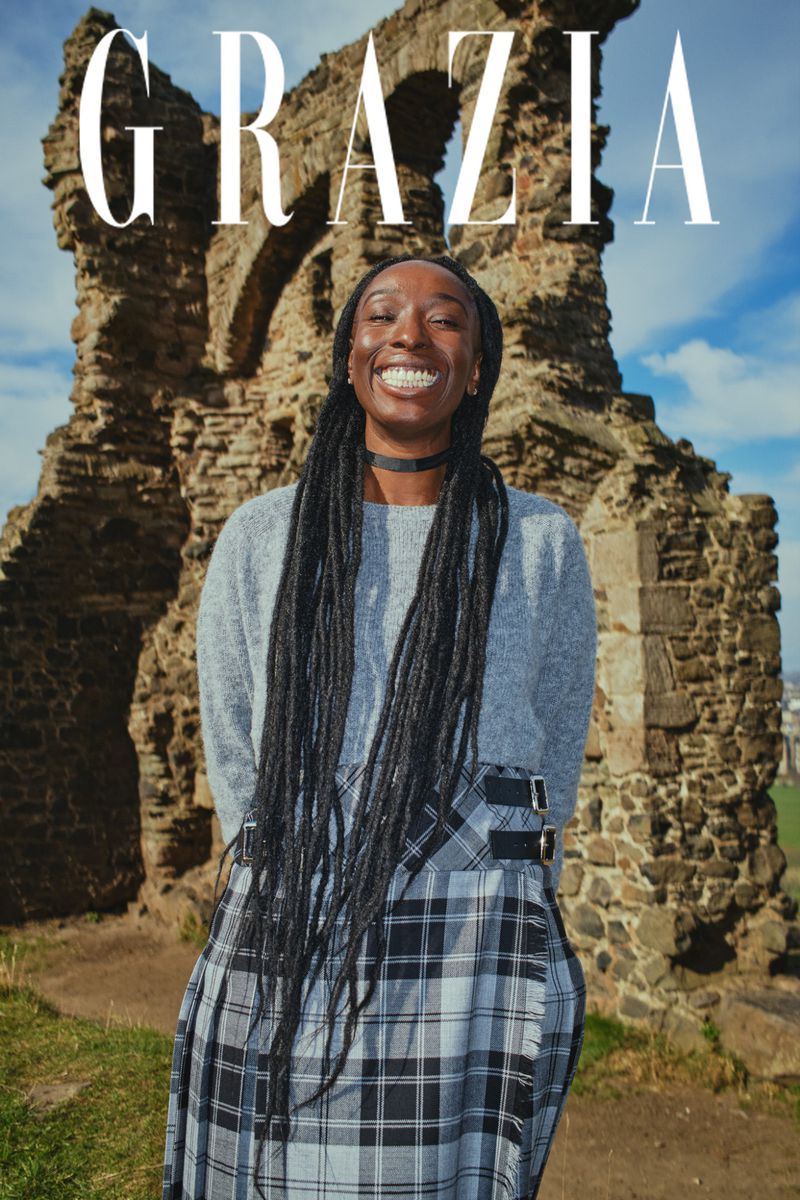 A smiling Black woman stands in front of ancient ruins, wearing a grey sweater and plaid skirt, under the magazine title GRAZIA.