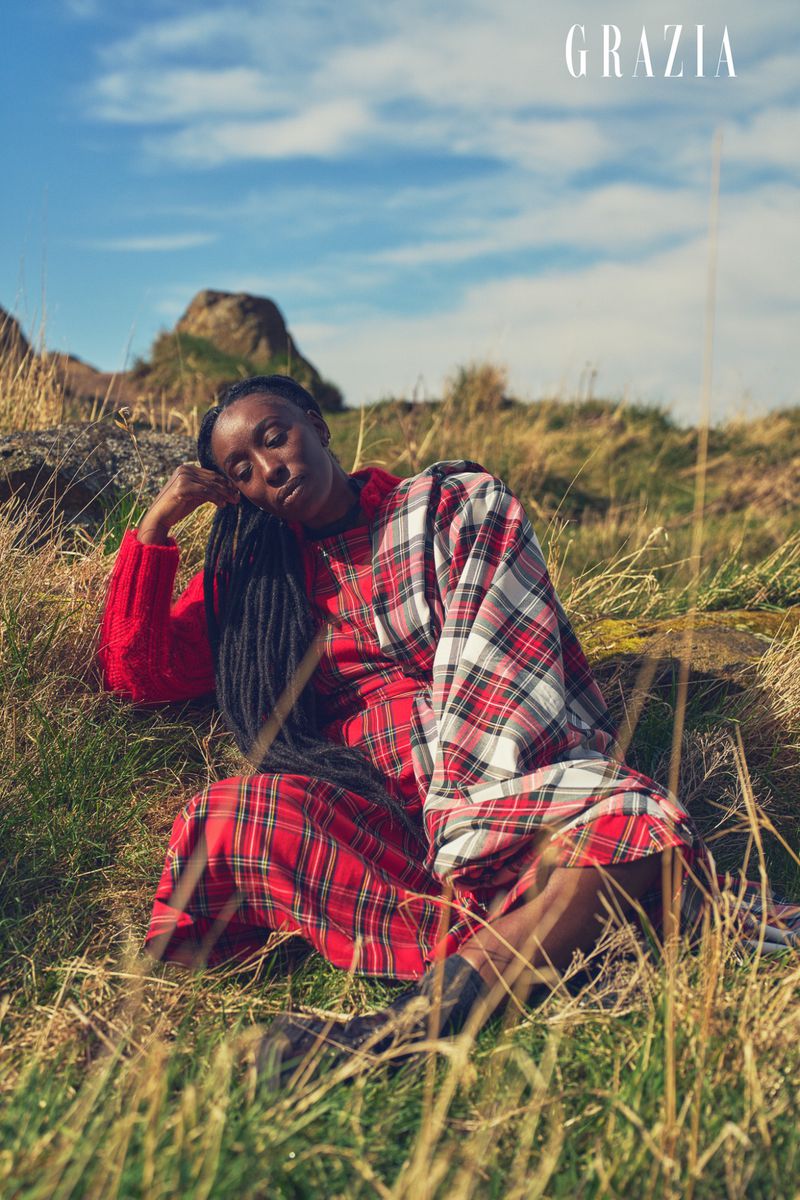 Woman in red plaid dress sits in a grassy field with rocky hills and blue sky, wearing a red sweater, and long braids.