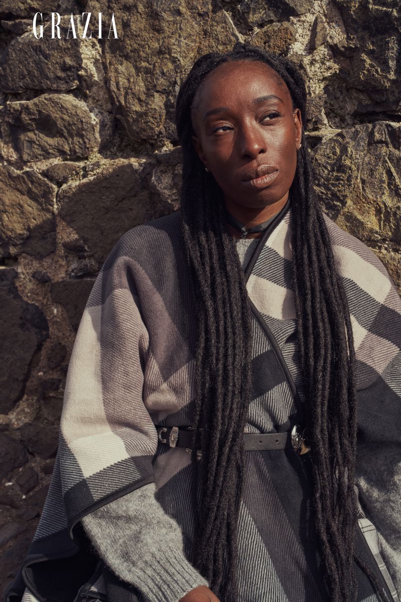 Portrait of an adult woman wearing a plaid poncho in front of a stone wall. She has dark skin and long black dreadlocks.