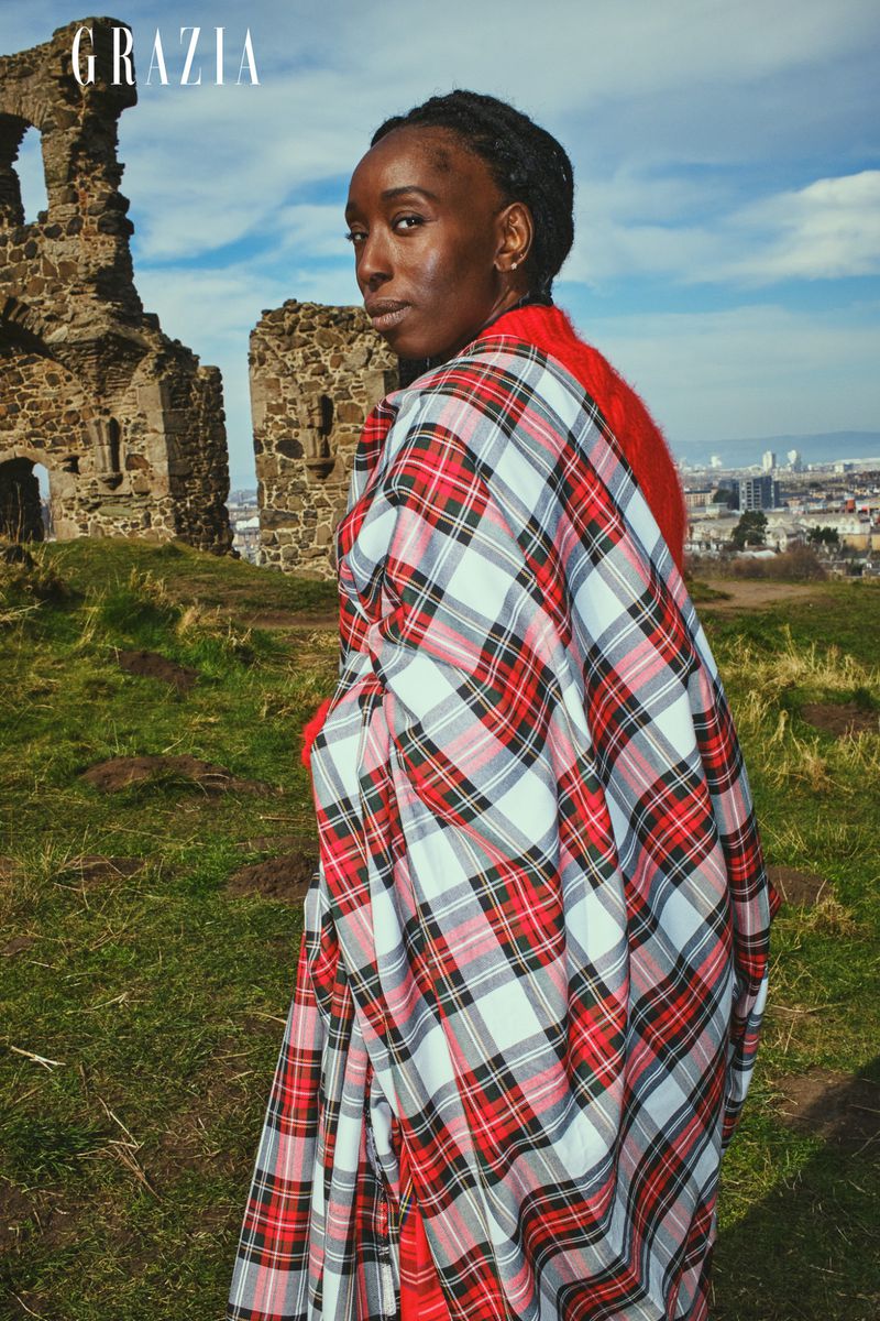 Woman in red and white plaid garment posing on grassy hill with stone ruin and city skyline in the background. Editorial fashion shoot.