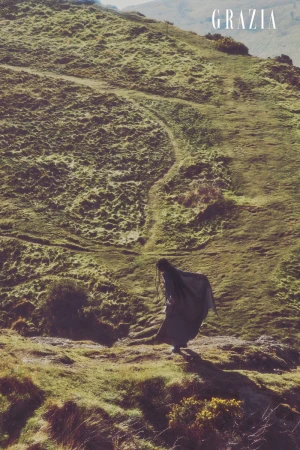 Model Eunice Olumide stands in a flowing dark cloak on a steep sunlit hillside landscape with a dirt trail.