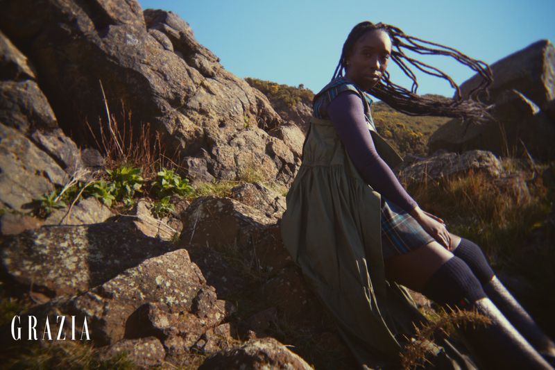 Outdoor fashion editorial featuring a Black female model in an olive dress, sitting on rocky terrain under a clear sky, style editorial.