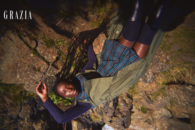 Fashion editorial style shot of an adult woman with braids wearing a plaid skirt and long sleeve shirt in an outdoor setting.