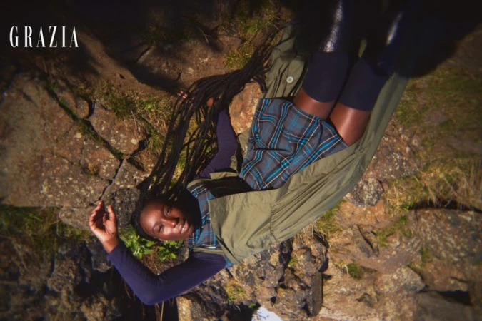 Fashion editorial style shot of an adult woman with braids wearing a plaid skirt and long sleeve shirt in an outdoor setting.