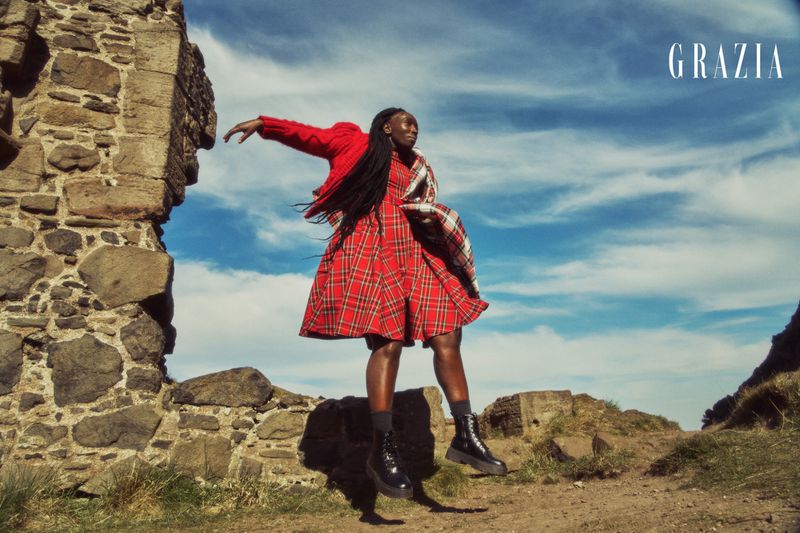 An adult female fashion model in a red plaid dress poses on a stone pathway with a stone wall and a cloudy blue sky in the background.