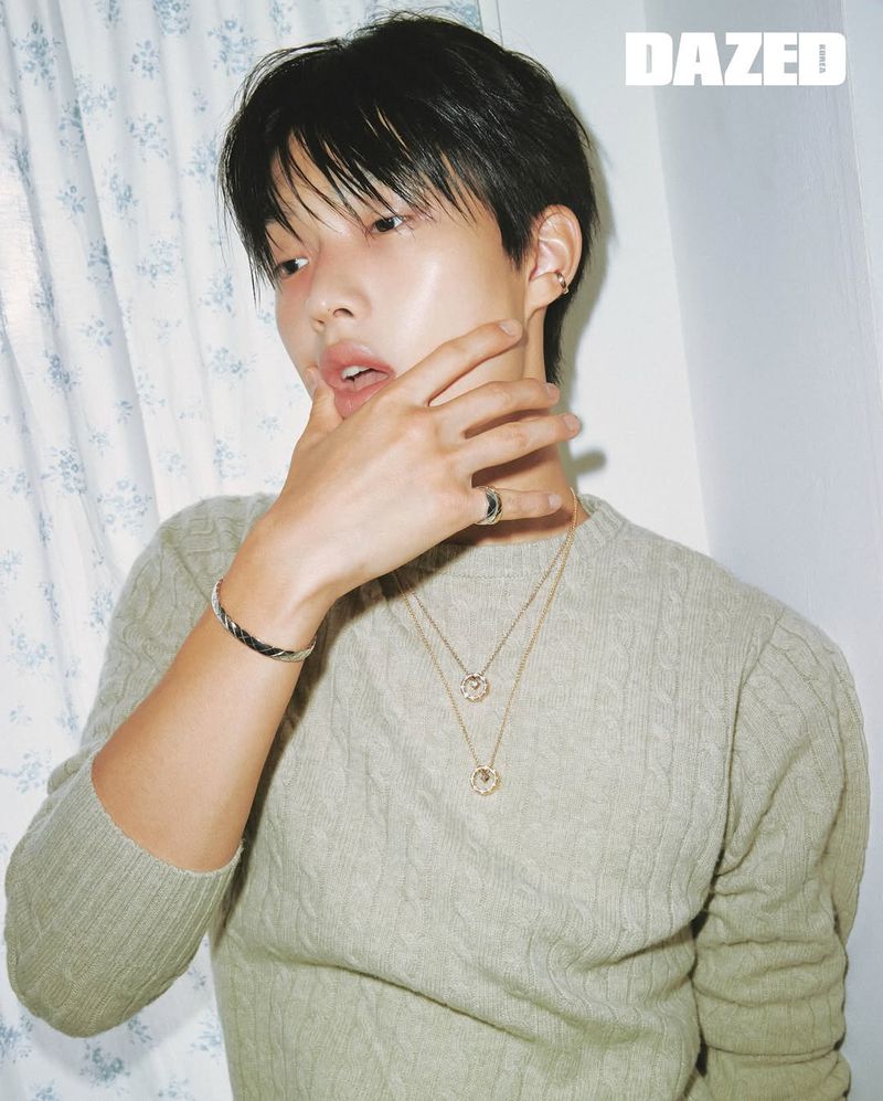 Close-up portrait of a young Asian male model with dark hair, wearing a knitted sweater and jewelry, against a white backdrop.
