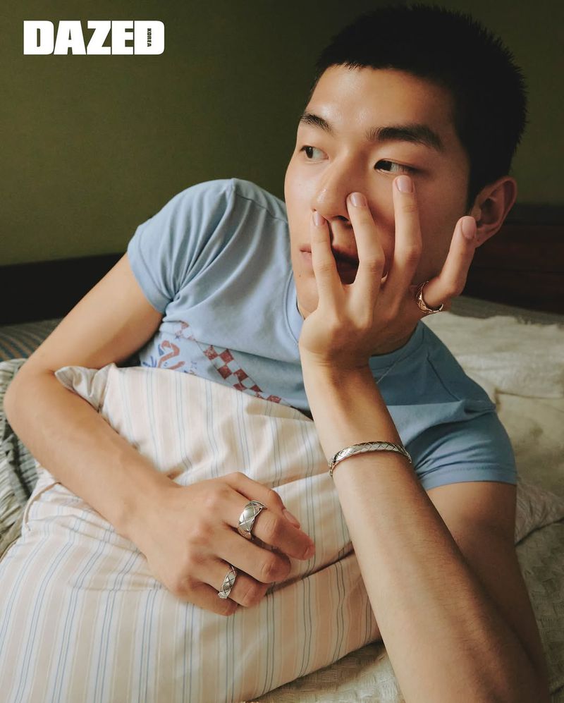 Portrait of an Asian adult male lying on a bed, with his hand near his face, wearing jewelry, and featuring a Dazed magazine logo.