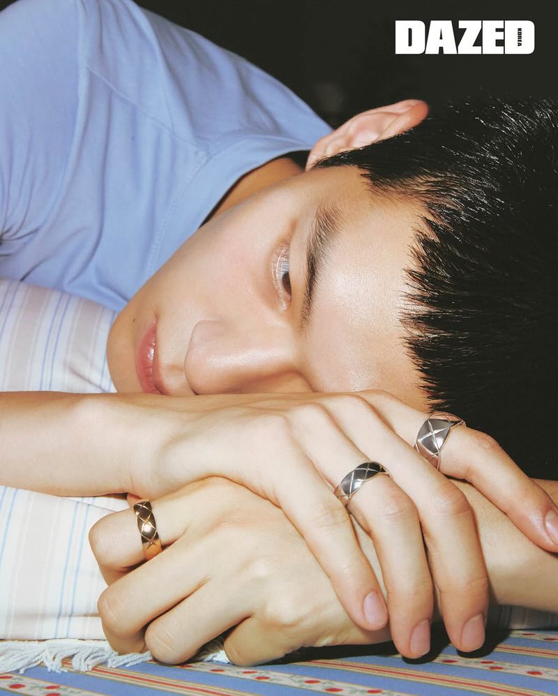 Close-up studio shot of an Asian male model with dark hair, wearing a blue top and rings, lying on a bed with a patterned bedsheet.
