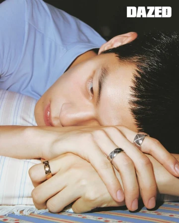 Close-up studio shot of an Asian male model with dark hair, wearing a blue top and rings, lying on a bed with a patterned bedsheet.