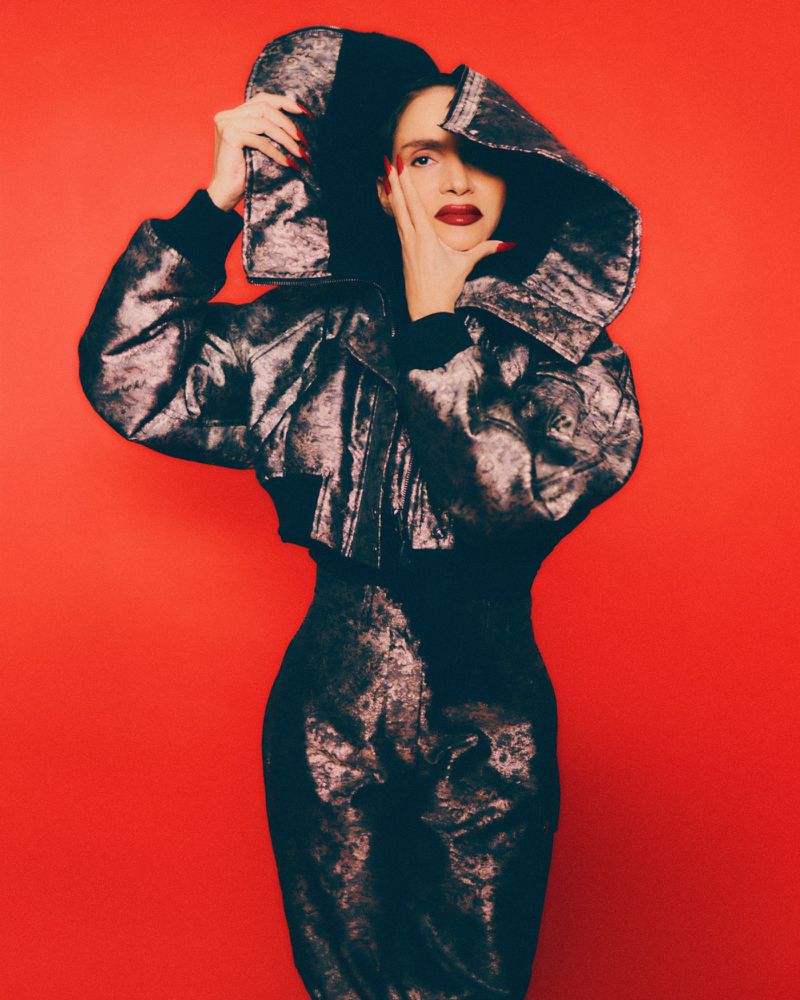 Woman in metallic jacket and matching skirt poses against a red backdrop, her hand near her face in a fashion forward portrait.