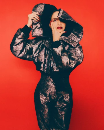 Woman in metallic jacket and matching skirt poses against a red backdrop, her hand near her face in a fashion forward portrait.