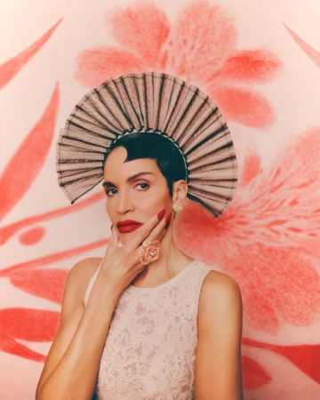 Elegant woman with floral-patterned dress and ornate headpiece poses in front of a pink floral backdrop, showcasing a unique style.