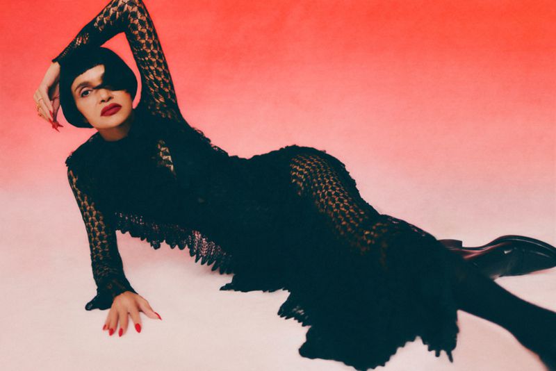 Woman in black lacy dress poses on a red gradient background. She's lying down, arm raised, and has red lipstick and nail polish.