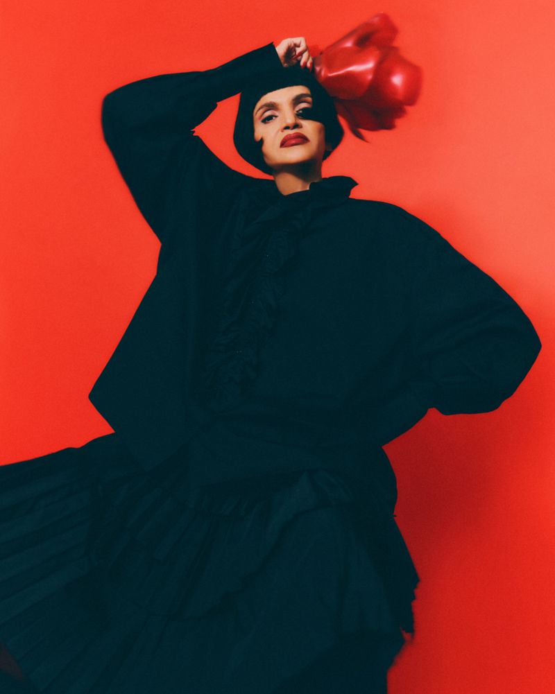 Adult female in black dress poses in front of a red background, holding red boxing gloves above her head in a studio setting.