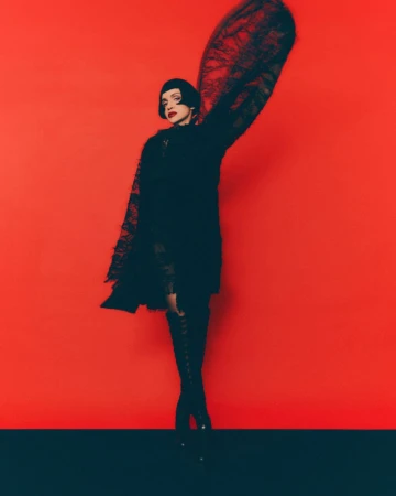 A woman with dark hair in a black dress and high boots poses against a red background with dramatic lace details and dark base.