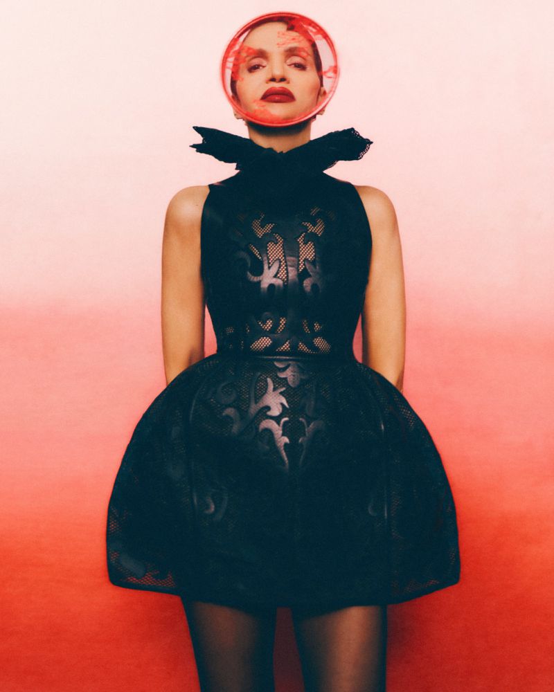 A woman in a black dress with a structured collar and a clear plastic head covering poses against a red gradient background in a studio shot.